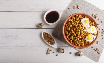 Fried chickpeas with quail eggs and spices in a clay plate on white wooden background, top view,