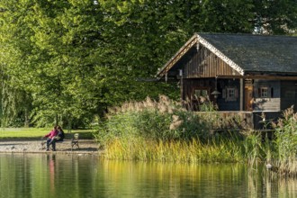 Wooden hut with common reed (Phragmites australis), two woman on a park bench, shore of Lake
