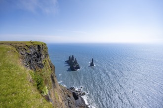 Young man standing on cliff, view from steep cliff to the rocks Reynisdrangar in the water, at