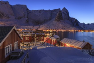 Red wooden houses on a fjord in front of snow-covered mountains, illuminated, twilight, winter,