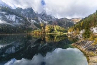 The Vordere Gosausee in autumn. The Gosaukamm on the left. Clouds and sun. Trees in autumn leaves.