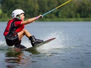 Boy or girl, sitting on wakeboard, cheerful, red life jacket, water skiing and summer holiday in