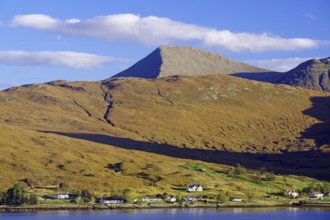 A tranquil inlet lies against a mountainous landscape of isolated houses and a blue sky, Cullins,