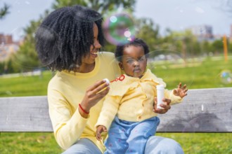 African girl on lap of her mother playing soap bubbles sitting on a park bench