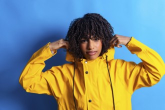 Studio shot of a young woman with curly hair adjusting the hood of her yellow raincoat against a