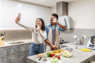 Young couple waving at the camera while preparing a healthy salad in their kitchen, making a video