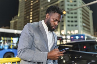 Young businessman using smartphone on city street at night, with blurred buildings and traffic in