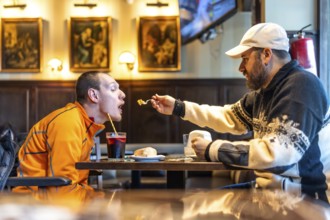 Man feeding his disabled friend in a restaurant, showcasing genuine care and inclusivity while