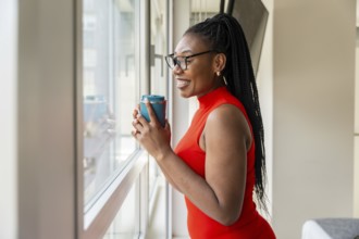 Smiling businesswoman drinking from a reusable cup while looking out the window of her hotel room