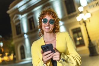Cool modern woman in sunglasses using mobile phone standing in the city at night