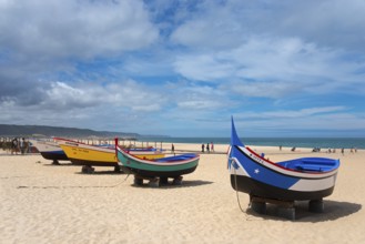 A row of colourful fishing boats on the sandy beach, some people walking along the shore, Praia da