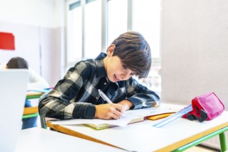 Young boy writing with pen on paper at desk in school classroom, smiling and focused during lesson