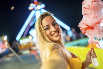 Woman taking selfie while eating pink cotton candy at fair during summer night