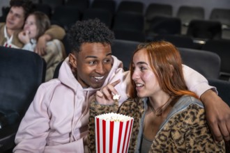 Happy diverse couple enjoying film and sharing popcorn bucket in movie theater, whispering during