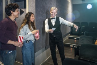 Young couple holding popcorn buckets while usher showing their seats in a movie theater, ready to