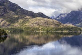 Calm ocean with clear reflection of forested mountains and cloudy skies in a peaceful setting,