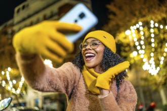 Smiling woman wearing yellow woolen gloves, hat and scarf taking selfie with her smartphone in a