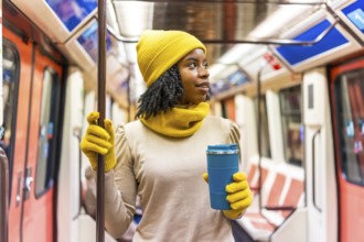 Black woman wearing yellow winter clothes holding a blue reusable coffee cup standing on a subway
