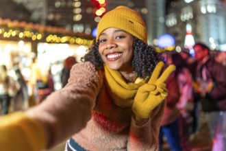 Smiling young woman wearing yellow winter clothes takes a selfie at christmas market showing peace