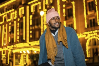 African man sitting outdoors during Christmas night facing Christmas fairy lights decorating a