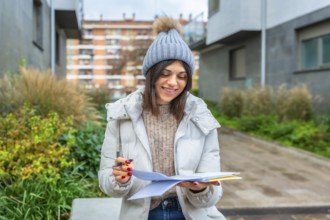 Smiling student writing notes in a notebook sitting on a city bench in winter