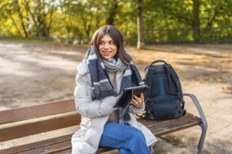 Woman using digital tablet sitting on a park bench during a sunny day of autumn