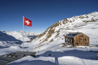 Swiss flag and mountain hut Keschhütte in front of mountain landscape in winter, behind summit of
