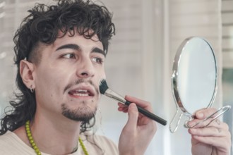 Young man with curly hair and piercings is applying makeup to his face using a brush while looking