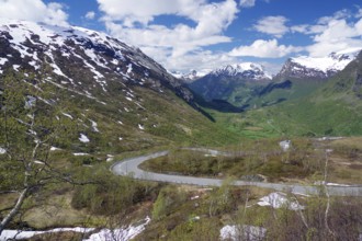 A winding road runs through a green valley between snow-capped mountains under a blue sky with