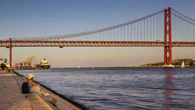A person sits by a riverbank, gazing at the bridge and boats during a calm sunset, 25 de Abril