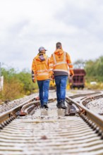 Two men in orange work clothes walking on rails, a wagon in the background, track construction