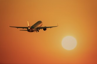 An aeroplane flies towards a large, orange glowing sun in the clear sky, take-off,