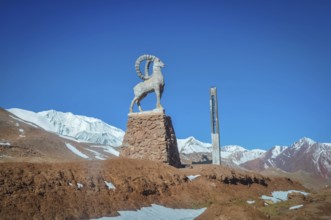 Sculpture of bouquetin on stony base surrounded by mountains with snowy peaks in distance in