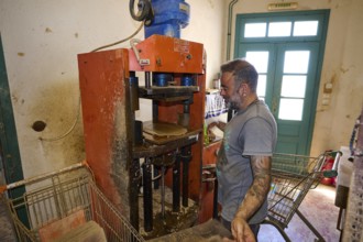 A man operates a brick press in a workshop, bricks and metal machine visible, Brickworks Patmos
