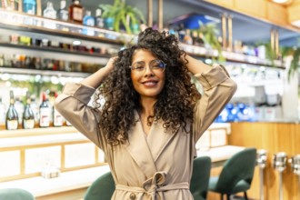 Sensual latin woman touching her curly hair and smiling at camera standing in a bar cafeteria
