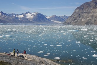People sitting on the shore with a view of a vast icy landscape and majestic mountains under a blue