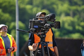 A cameraman in a brightly coloured waistcoat with a camera films outside in sunny weather against a