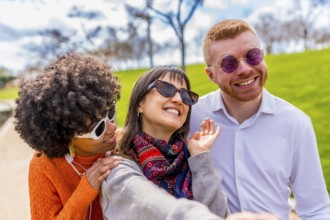 Three cheerful friends, wearing sunglasses, are taking a selfie in a park, enjoying a sunny day and