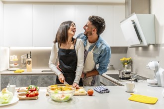 Young loving couple wearing aprons, preparing a healthy salad together in their modern kitchen,