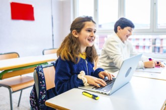 Smiling elementary school girl using laptop during lesson in classroom, next to classmate writing