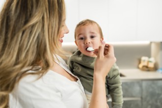 Smiling mother feeding yogurt to her baby son with a spoon in a bright kitchen, healthy eating