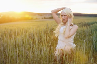 A blonde woman in a white dress sits in a grassy field under soft sunset light