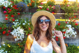 A woman wearing a straw hat and sunglasses is smiling in front of a wall of flowers. The flowers