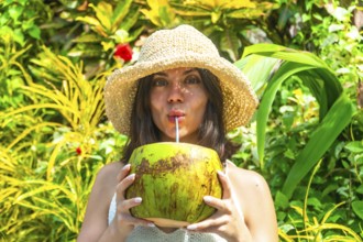 Young woman with straw hat savoring refreshing coconut water, surrounded by vibrant tropical