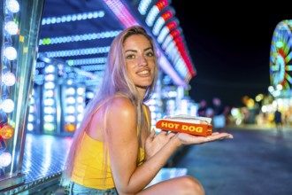 Blonde skinny woman eating a hot dog in an amusement park at night