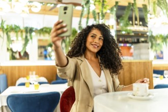 Happy and beauty latin woman with long curly hair and chic clothes taking selfie sitting alone in
