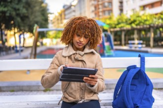 Latin beauty female student using digital tablet sitting on an urban park bench in the city