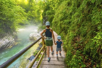 Mother and son wearing helmets are walking on a wooden pathway alongside a pristine river, enjoying