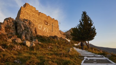Warm evening light, stone building with OXI writing, next to it a tree, person on the way,