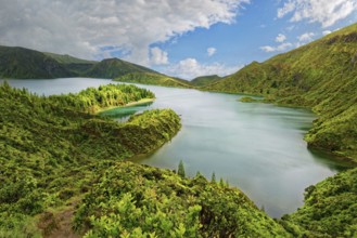 A tranquil lake surrounded by green mountains and wooded slopes under a blue sky, crater lake Lagoa